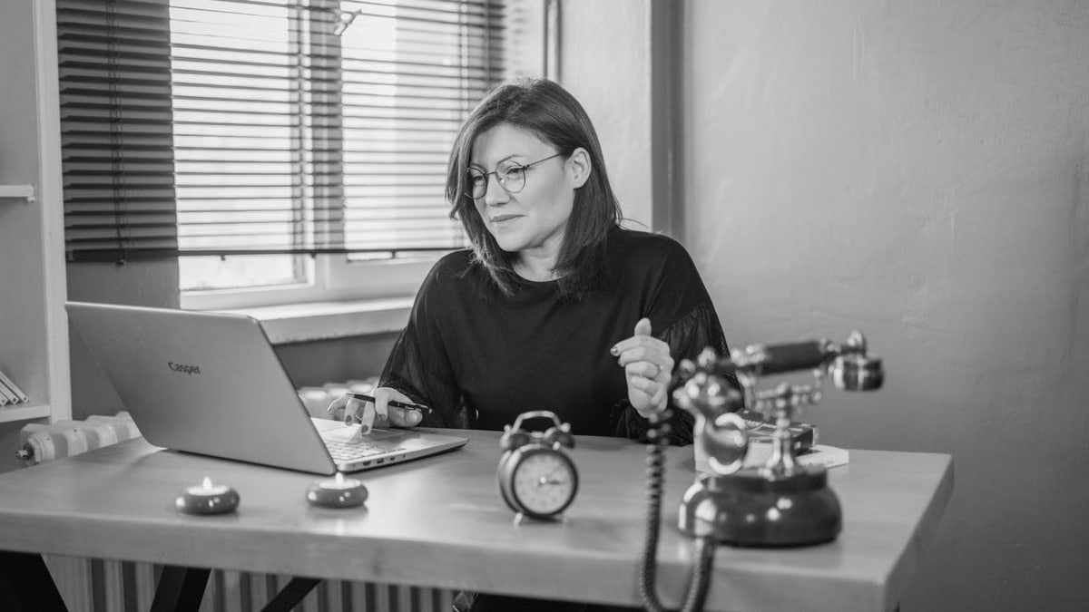 Black and white photo of a woman working at a desk with a laptop and vintage telephone.