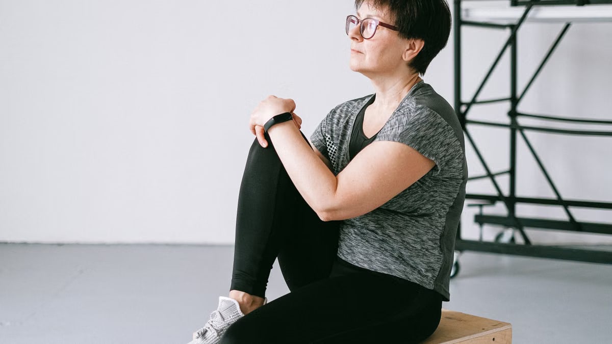 Senior woman in active wear sitting on a wooden box indoors, reflecting on wellness.