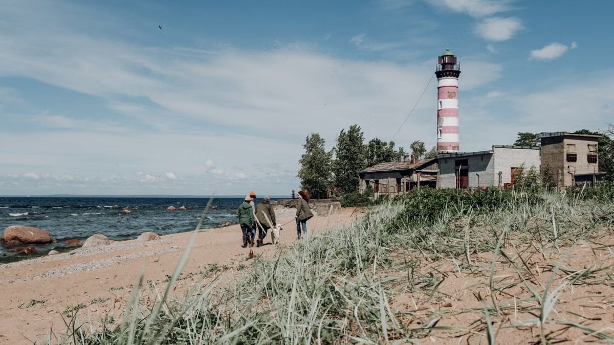 A group of people and a dog walk along a sandy beach near a lighthouse under a blue sky.